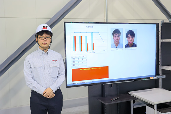 鹿児島姶良工場 事務の菱刈達矢。担当は事務がメインだが、フロントのサポートも行っている。写真のモニターは各班の作業の進捗状況を見える化するもので、菱刈がリーダーとなって導入した